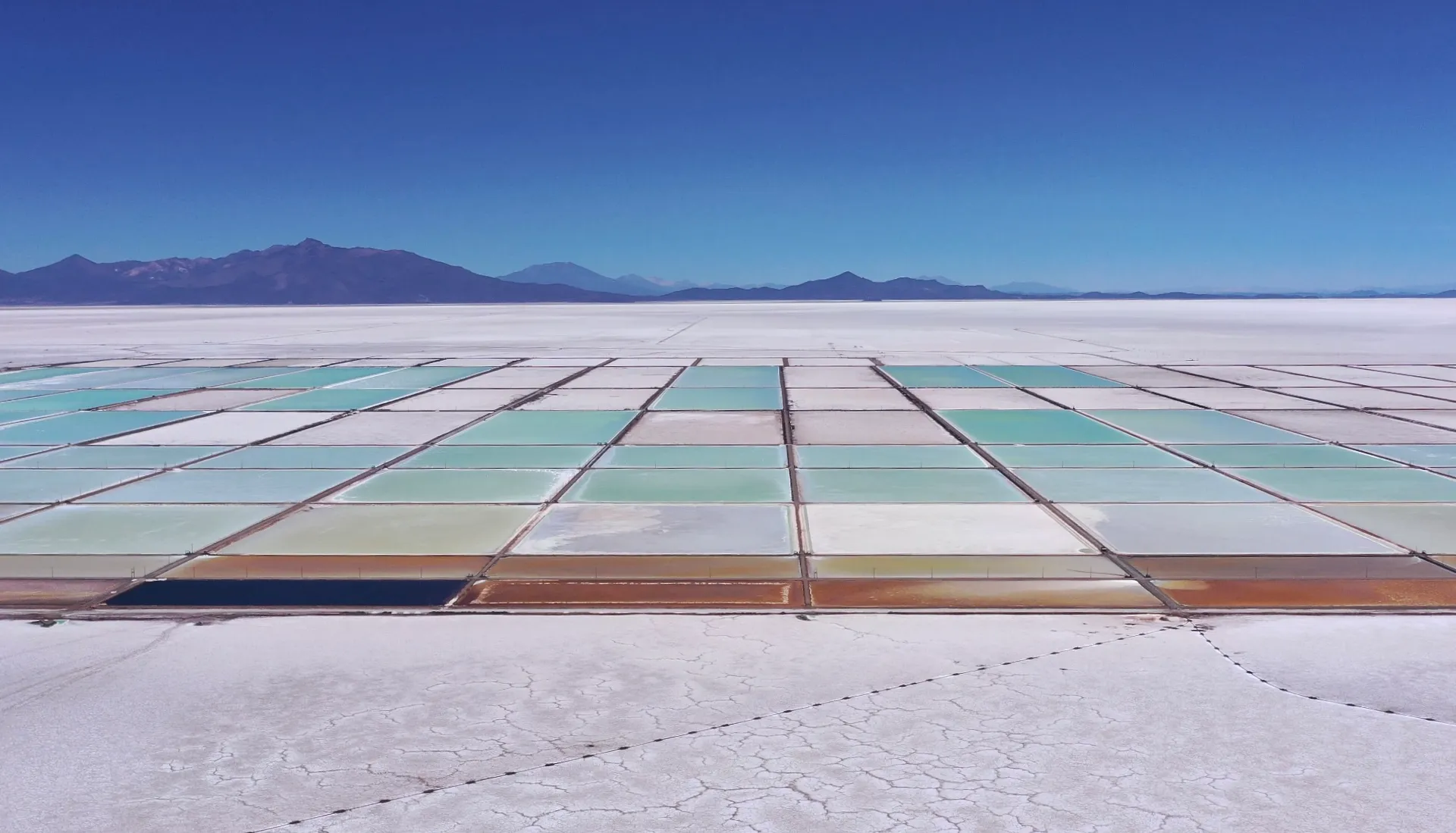 Vast white salt flat with colorful mineral evaporation ponds and distant mountains.